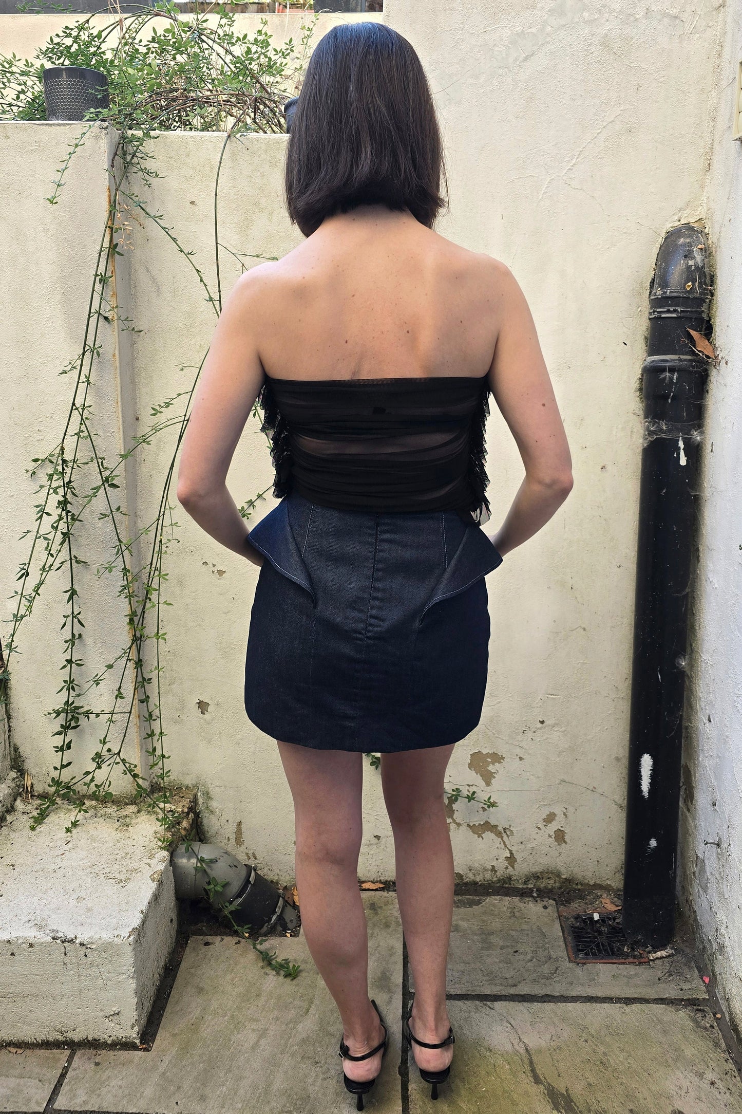 A woman viewed from behind wearing a strapless black top with sheer mesh panels and fringe, paired with a dark denim mini skirt, standing outdoors against a textured cream wall.