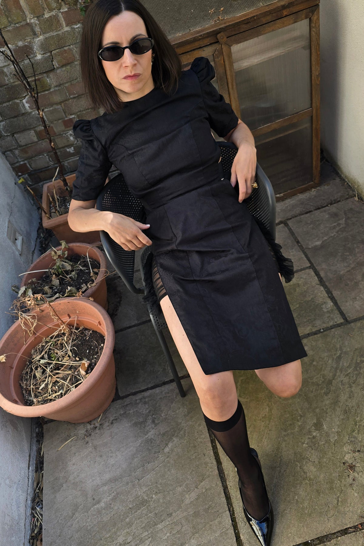 A woman wearing a black puff-sleeve midi dress, black thigh-high socks, and black pointed-toe heels sits on a chair in an outdoor patio setting.
