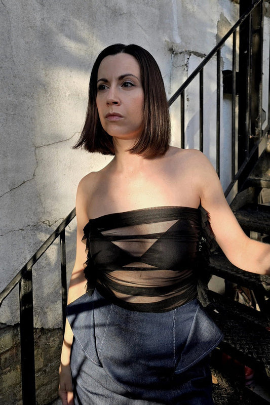 A woman with shoulder-length dark brown hair stands on outdoor stairs wearing a strapless black bandeau top with sheer crisscross mesh panels and blue denim bottoms.