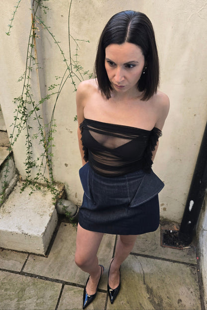 A woman with dark hair stands outdoors wearing a sheer black strapless top with a dark denim mini skirt and black pointed heels.