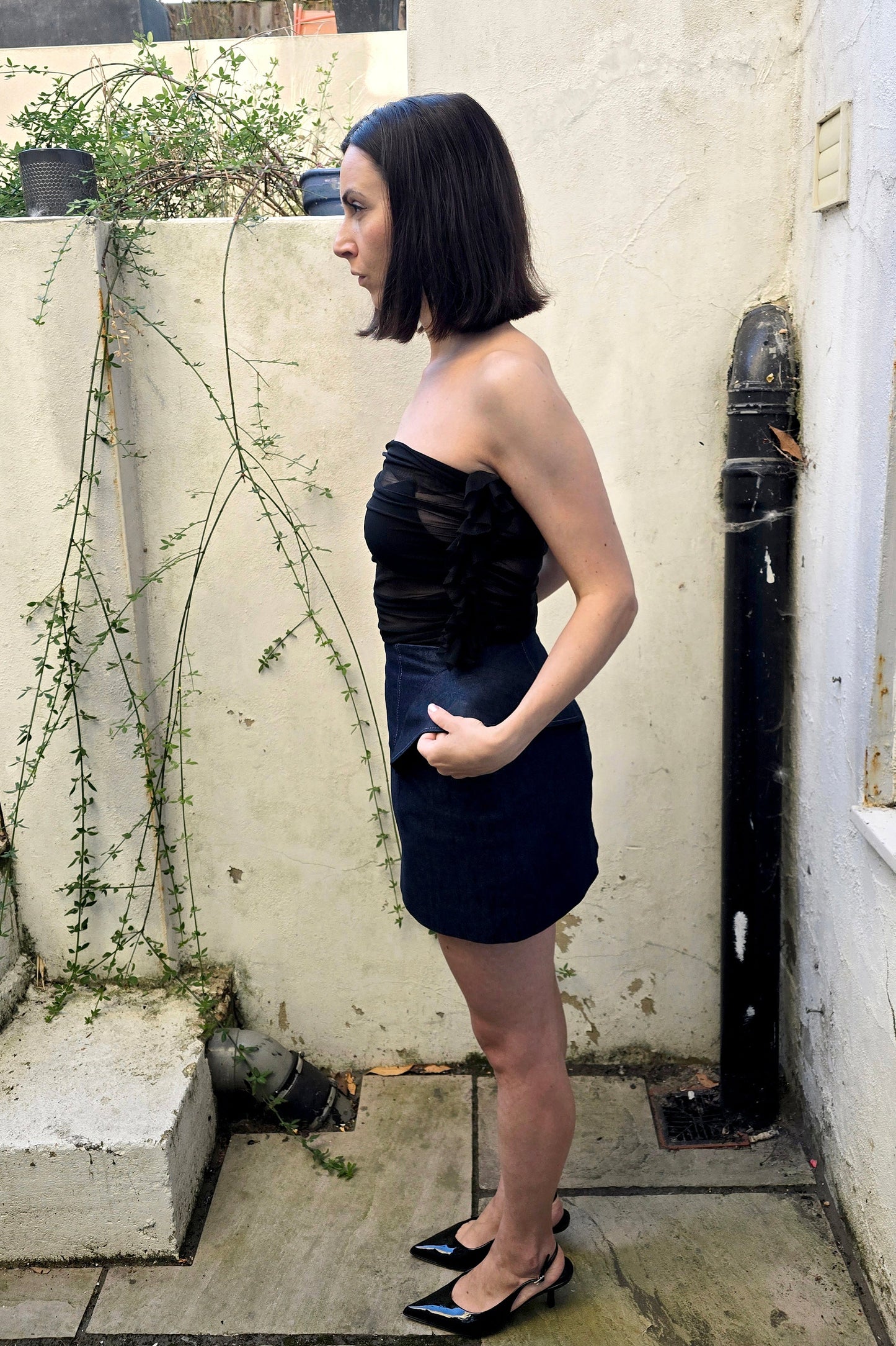 A woman with a dark bob haircut stands in profile wearing a strapless black mini dress and black pointed slingback heels, posing outdoors against a beige stucco wall.