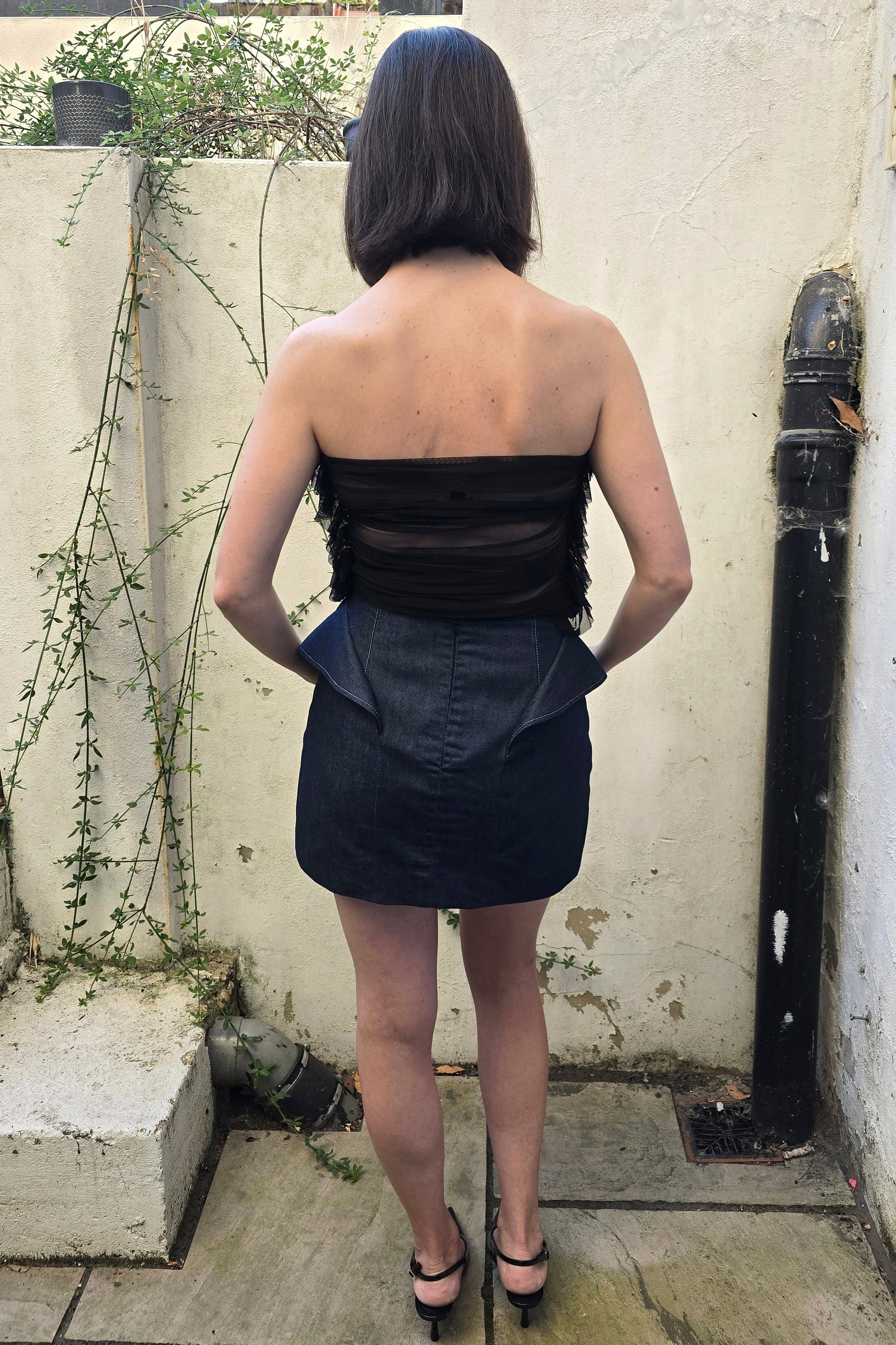 A woman viewed from behind wearing a strapless black top with sheer mesh panels and fringe, paired with a dark denim mini skirt, standing outdoors against a textured cream wall.