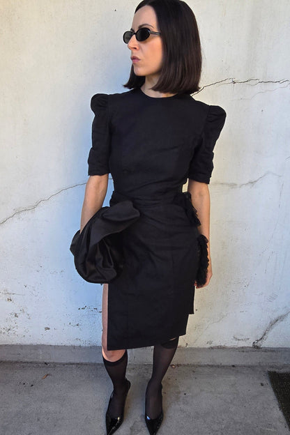 A woman with short dark hair and sunglasses wearing a black puff-sleeve dress with a tied waist, black tights, and black pointed shoes, standing against a textured off-white wall.