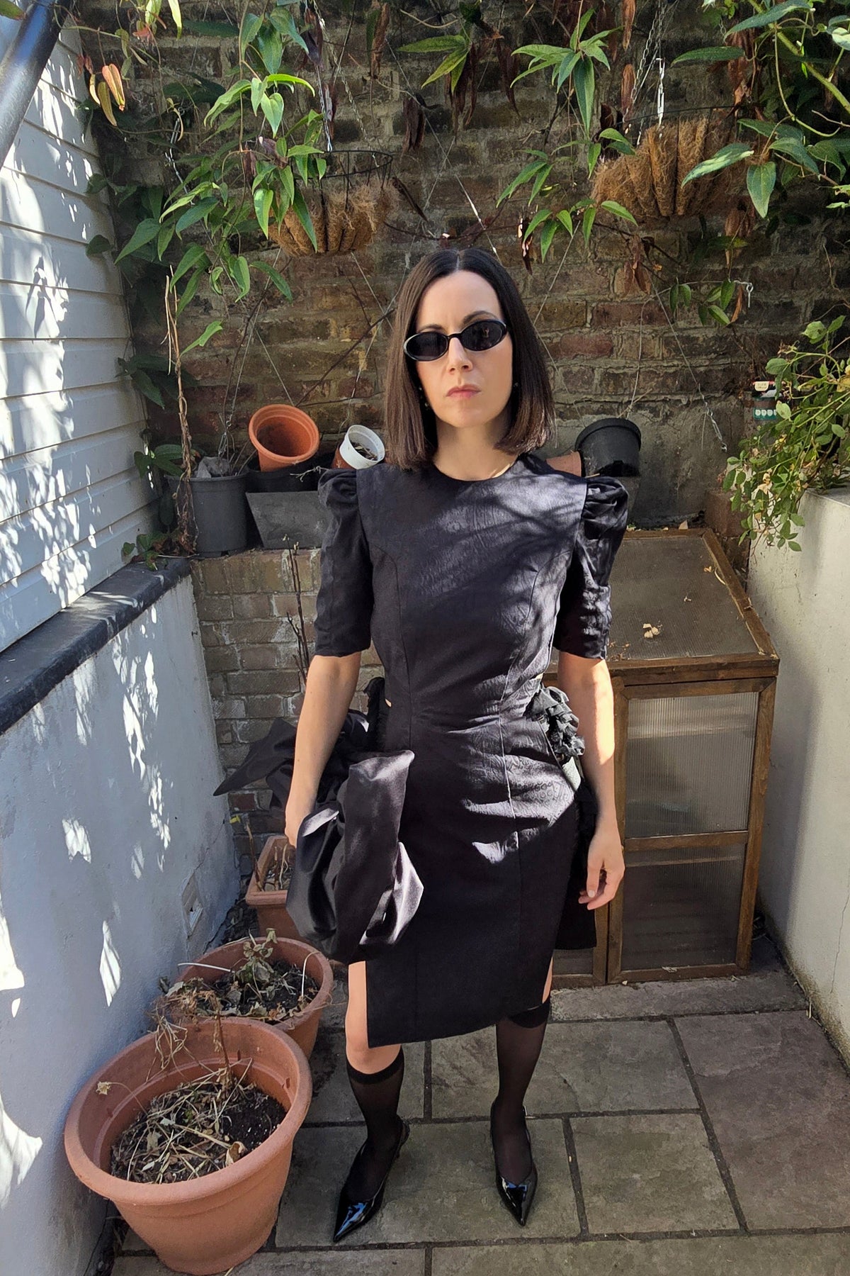 A woman stands on a stone patio wearing a black short-sleeve dress with structured shoulders, black tights, and black pointed shoes. She is posed in front of a brick wall with potted plants and a small wooden table.