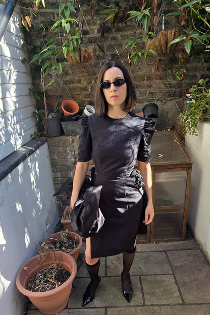 A woman stands on a stone patio wearing a black short-sleeve dress with structured shoulders, black tights, and black pointed shoes. She is posed in front of a brick wall with potted plants and a small wooden table.