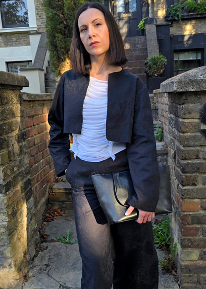 A woman stands outdoors on a stone path between brick walls, wearing a black cropped jacket over a white top, black wide-leg trousers, and black boots, while holding a metallic clutch bag.