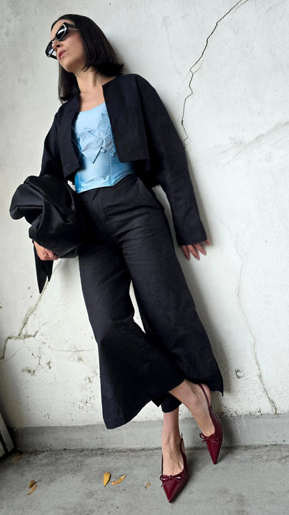 A woman posing against a cracked concrete wall wearing a black cropped Nicole Quadrio jacket, black wide-leg pants, a light blue top, and burgundy pointed-toe heels.