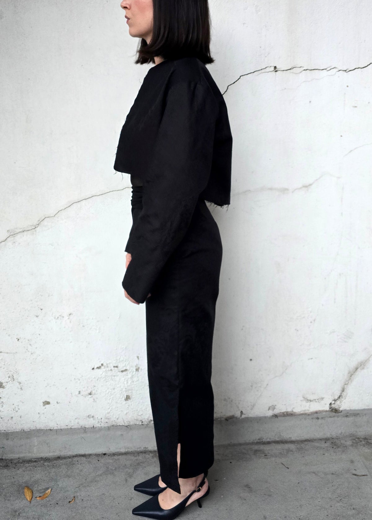 A woman stands in profile against a textured white wall, wearing a black long-sleeve top with dramatic draped shoulders and matching black trousers, paired with black slingback heels.