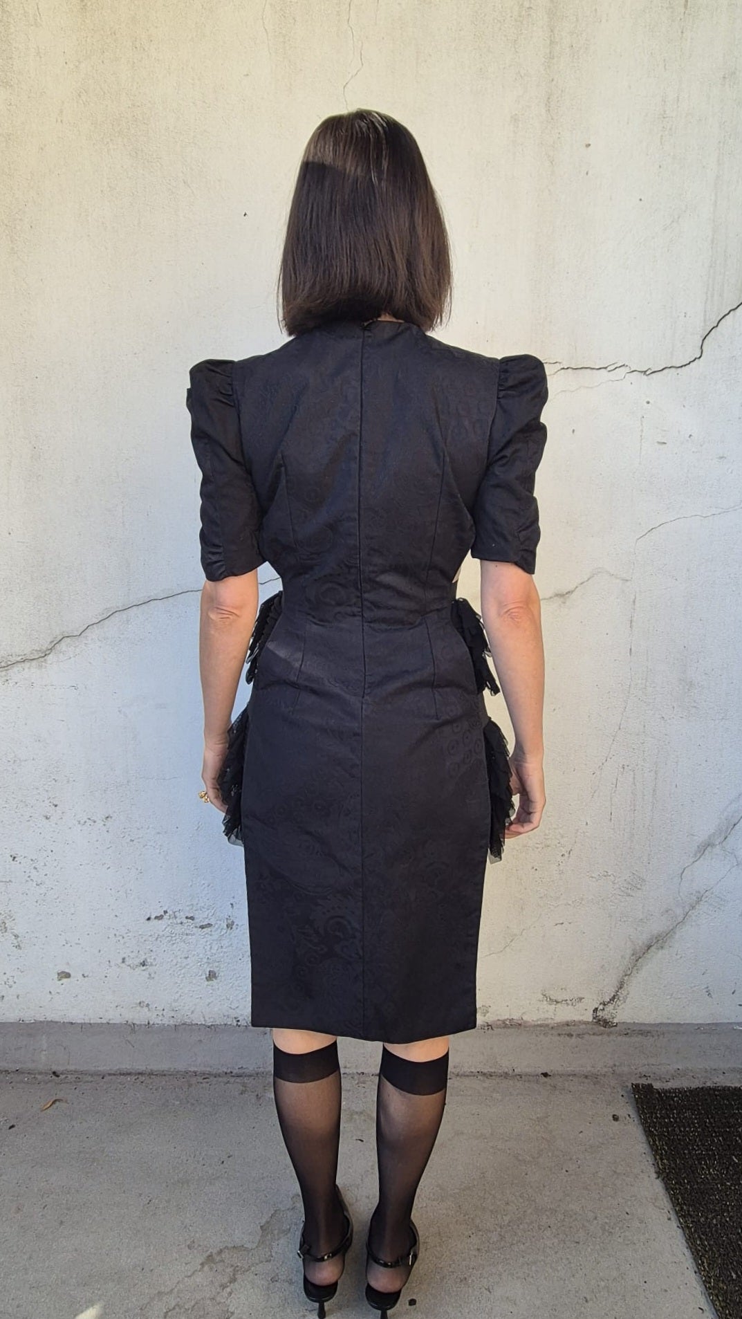 A woman viewed from behind wearing a black puff-sleeve pencil dress with a structured waist, paired with sheer black tights and black heels, standing against a textured off-white concrete wall.