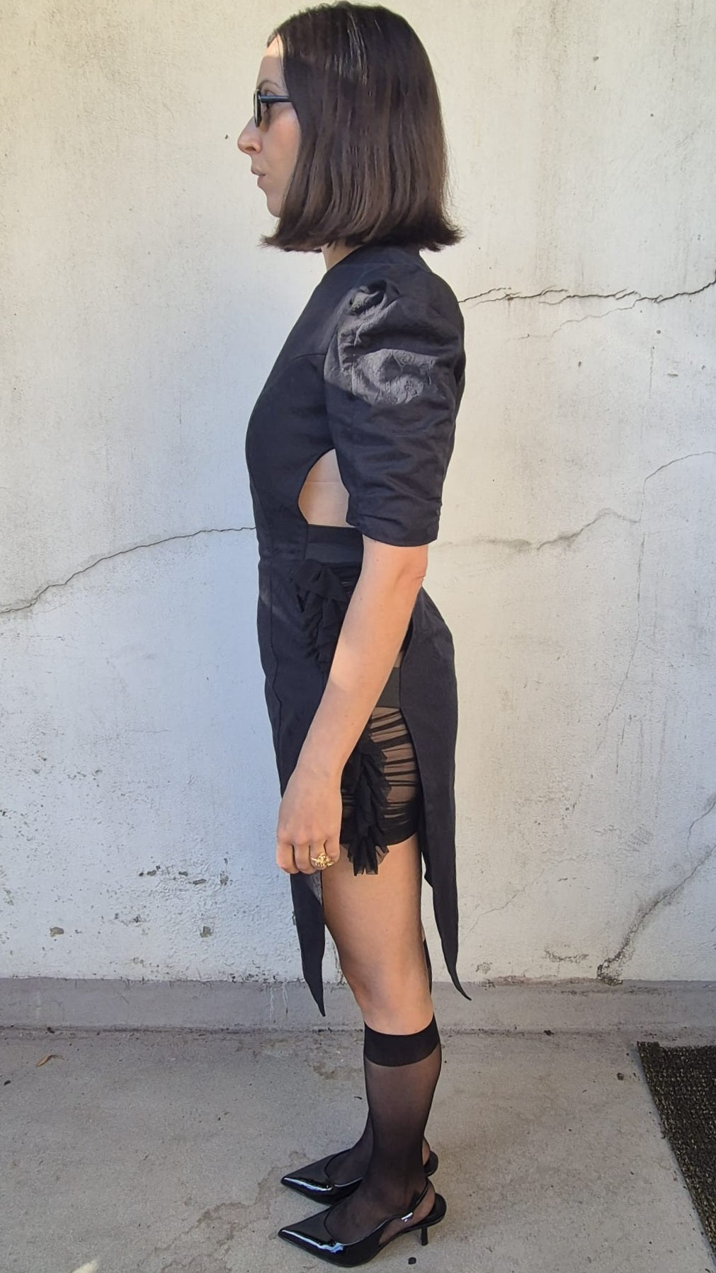 A woman in profile wearing a black short-sleeve dress with textured puff sleeves, side cutouts, and fringe details, paired with sheer black knee-high socks and black pointed-toe flats, standing against a textured concrete wall.