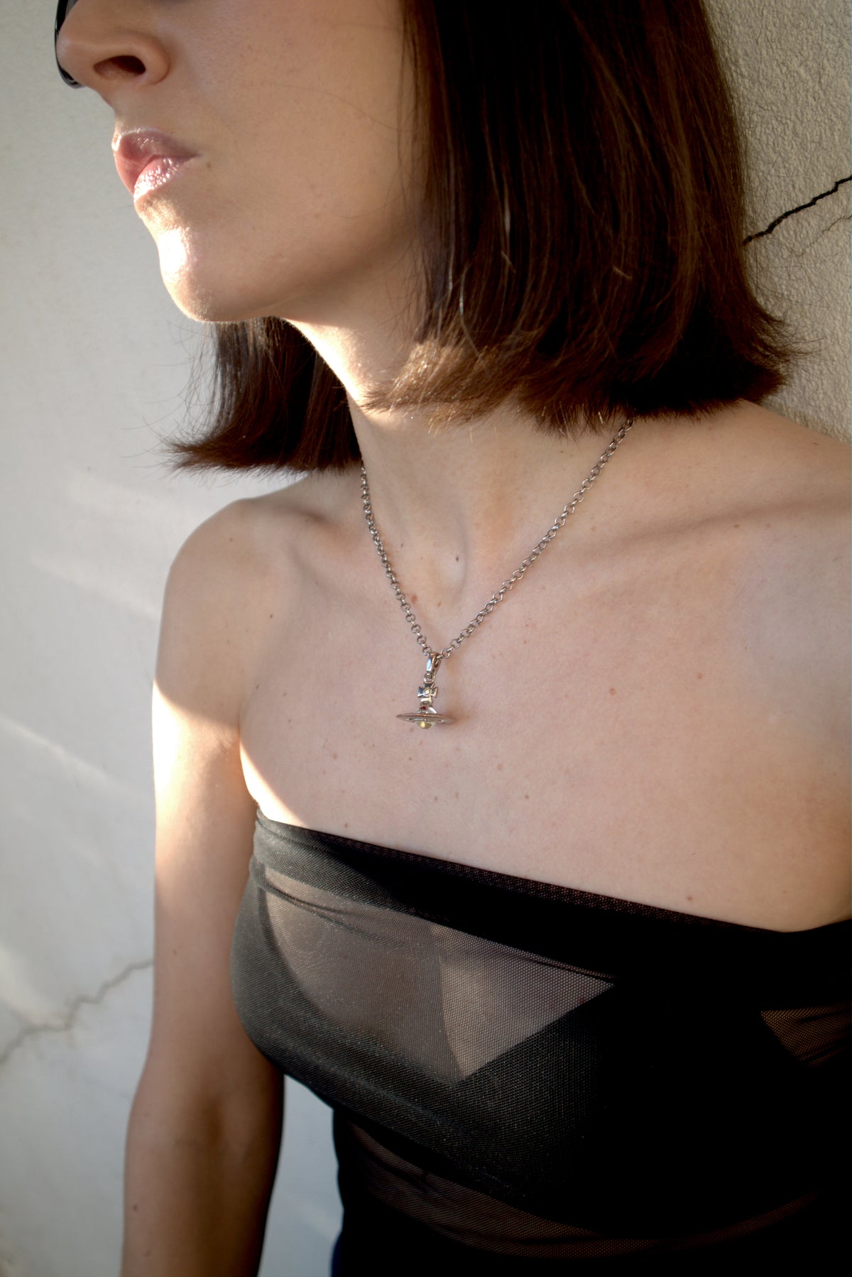 A close-up lifestyle shot of a woman wearing a silver-toned chain necklace with a small cross pendant, paired with a black strapless top against a textured off-white wall.